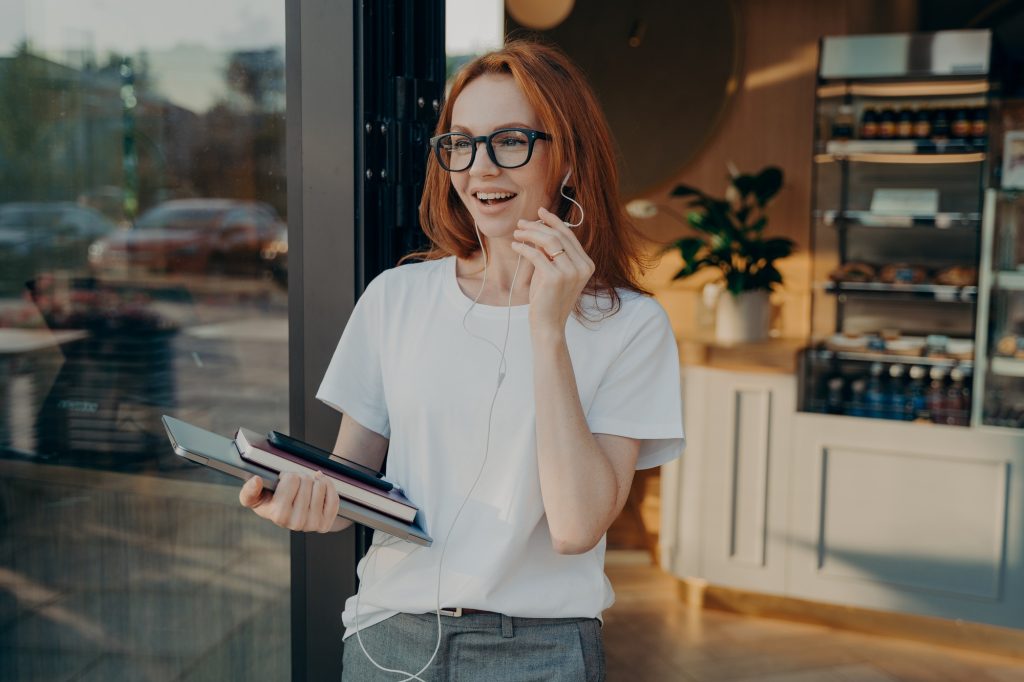 Happy red-haired businesswoman with laptop in hand having call on mobile phone, leaving coffee shop